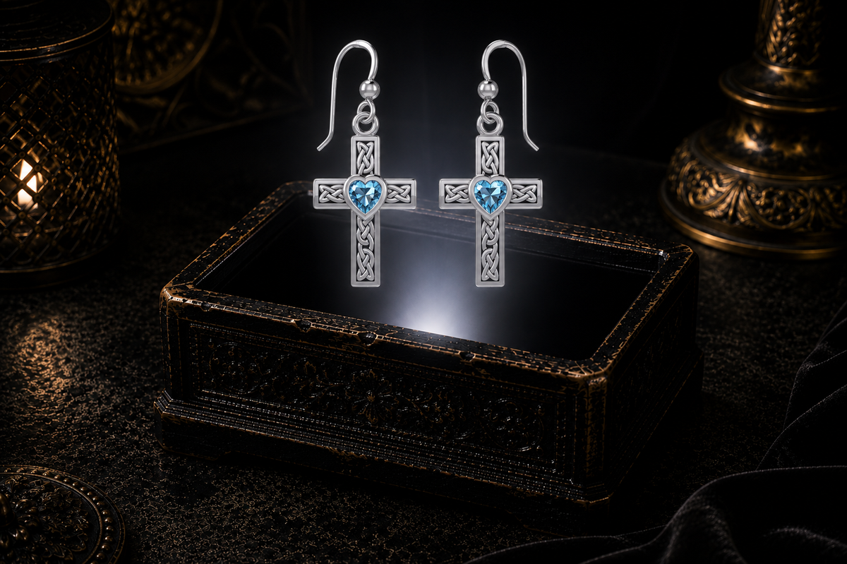 Sterling silver Celtic cross earrings with blue topaz heart gemstones floating above a glowing ornate jewelry box in a dark luxury setting