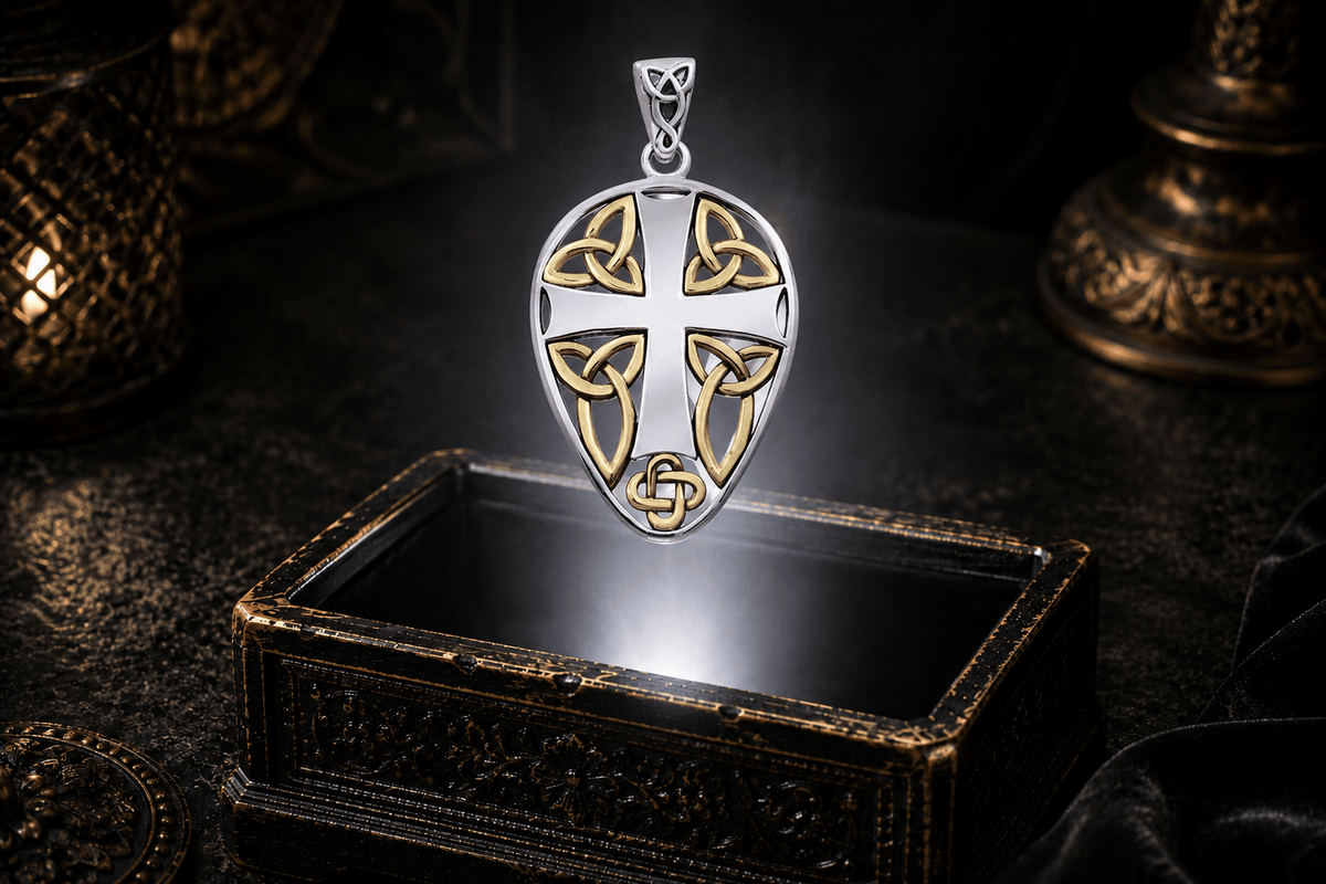 Sterling silver Celtic cross shield pendant with gold knotwork accents suspended above an antique jewelry box in a neutral white beam of light.