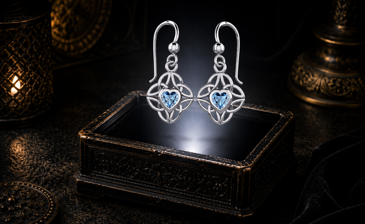 Sterling silver Celtic heart earrings with four interconnected Triquetra knots and blue topaz gemstones floating above an ornate jewelry box in a dark luxury setting with mystical glowing light.