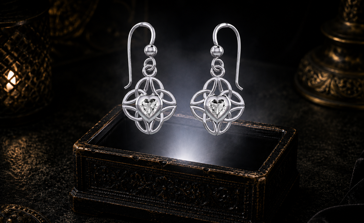 Sterling silver Celtic heart earrings with four interconnected Triquetra knots and cubic zirconia gemstones floating above an ornate jewelry box in a refined dark luxury setting with luminous light.