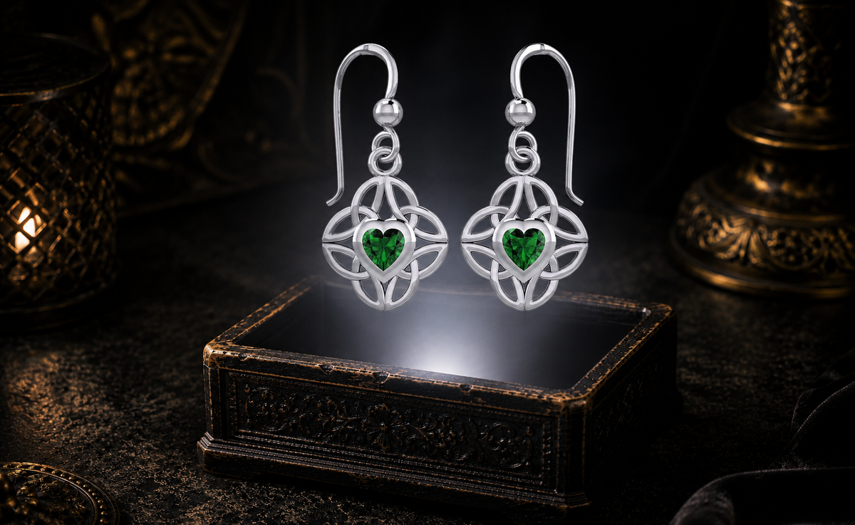Sterling silver Celtic heart earrings with four interconnected Triquetra knots and emerald glass gemstones floating above an ornate jewelry box in a cinematic dark luxury setting with glowing light.