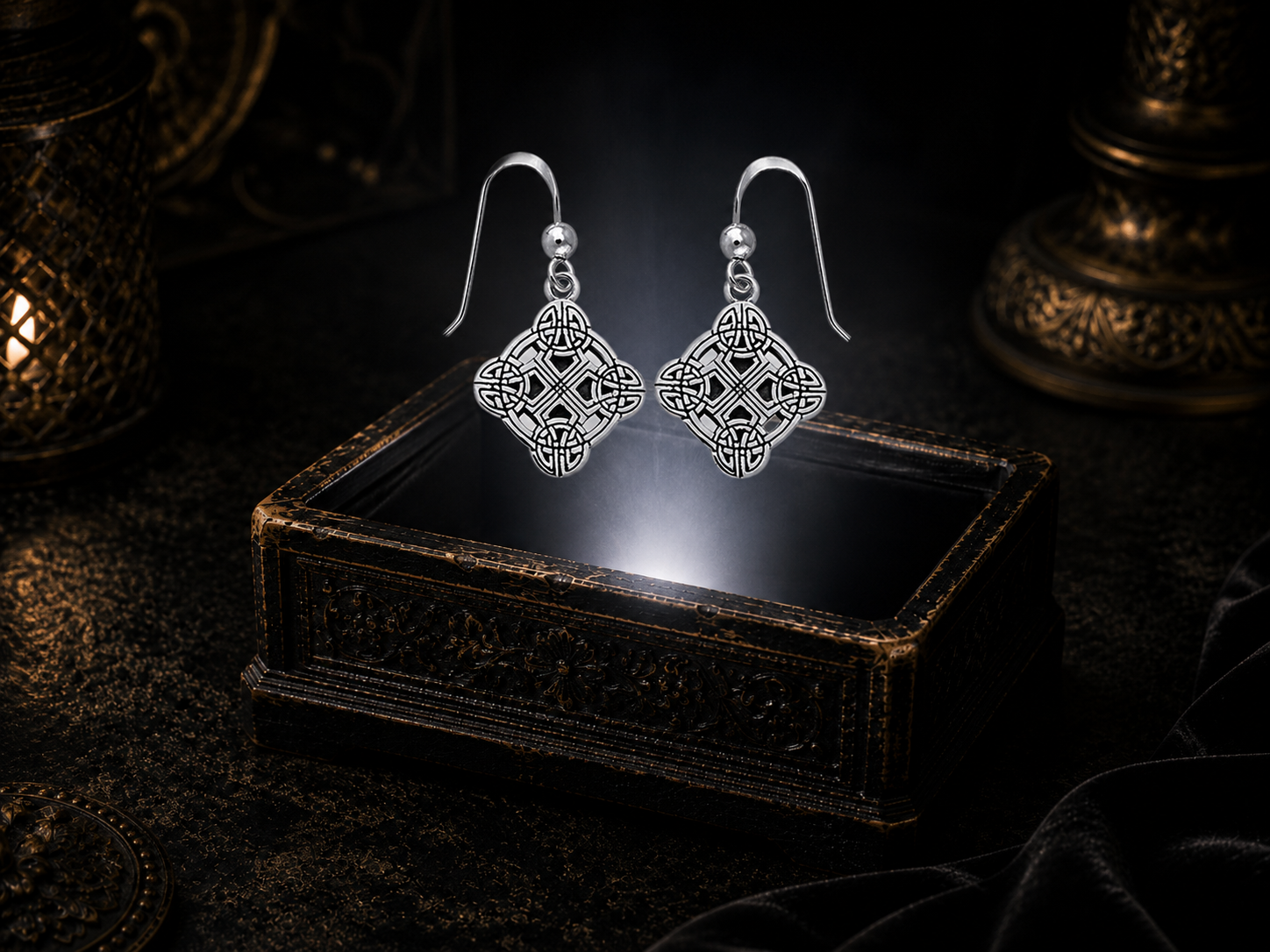 Sterling silver Clonmacnoise Celtic cross drop earrings floating above a glowing ornate jewelry box in a dark luxury setting