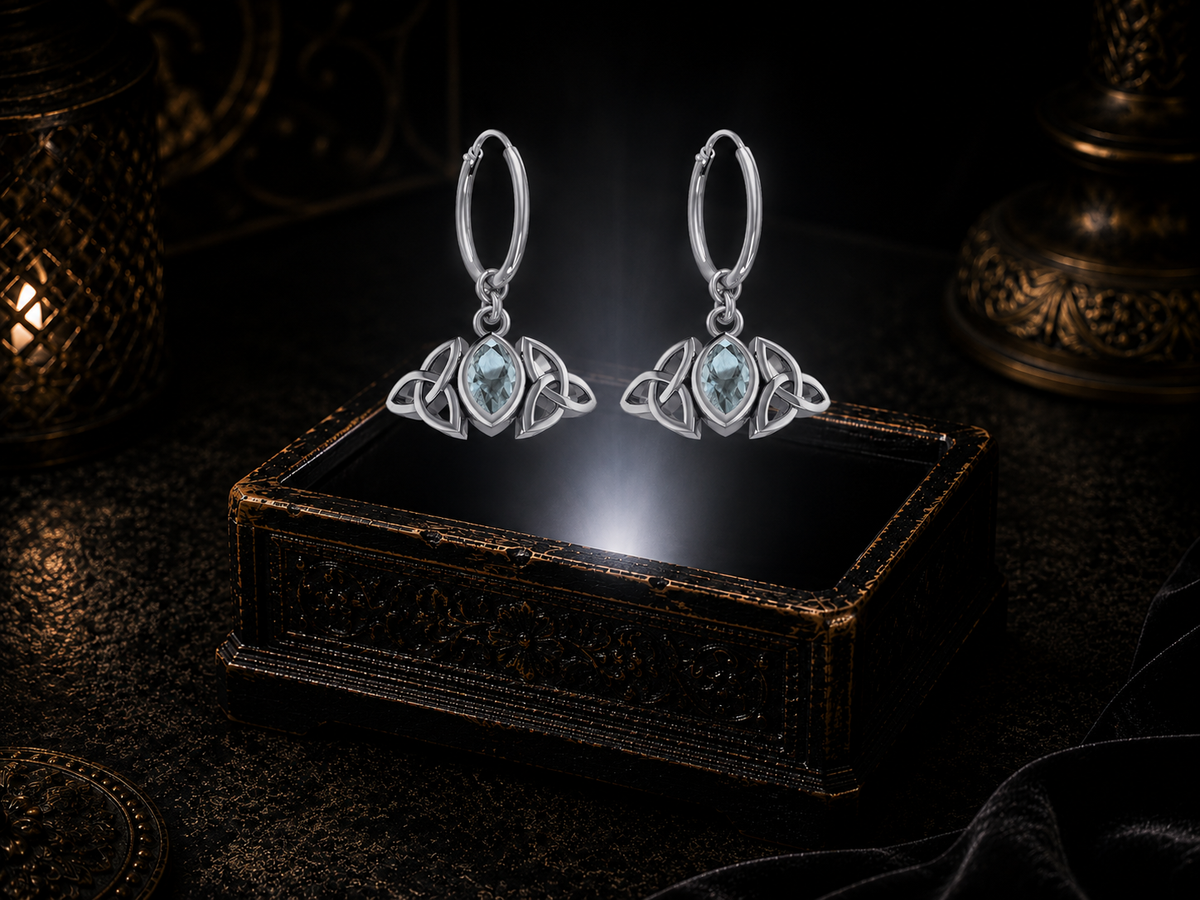Sterling silver Triquetra charm hoop earrings with blue topaz gemstones floating above a glowing ornate jewelry box in a dark luxury setting